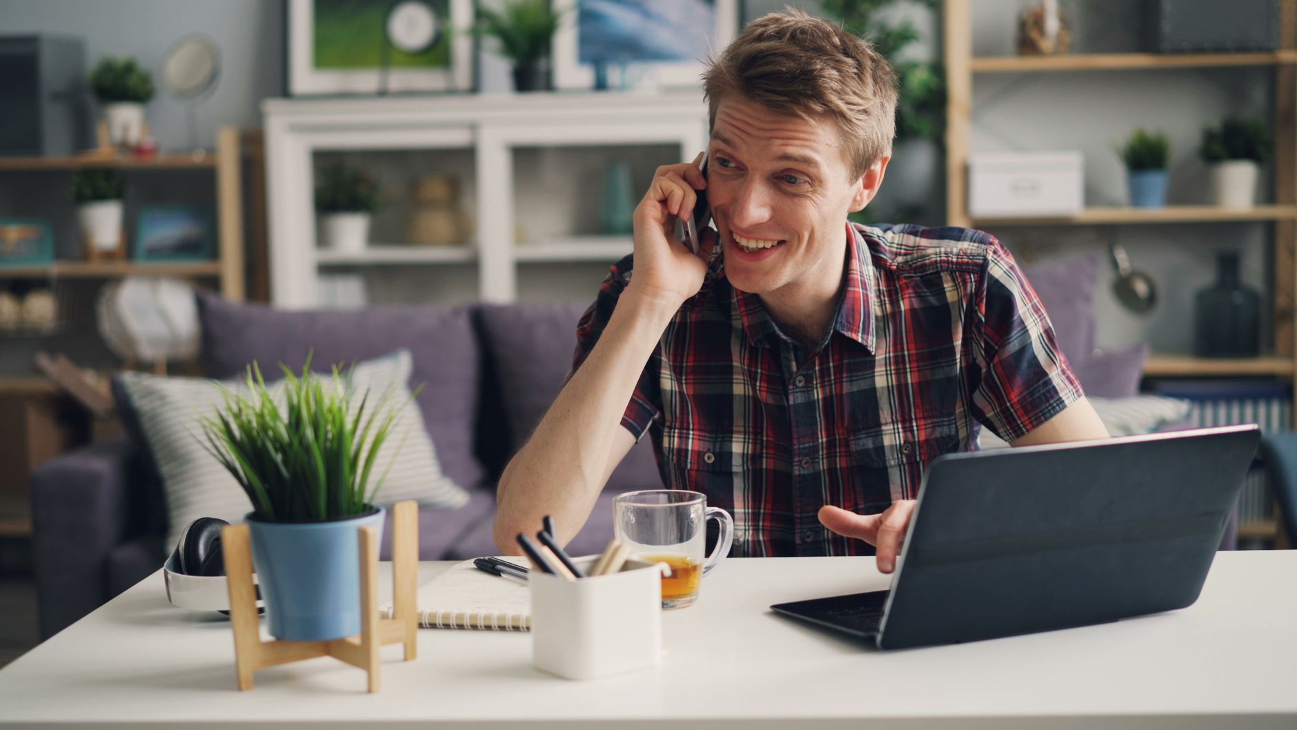 Mann i rutete skjorte snakker i telefonen på hjemmekontoret, med bærbar PC og kaffekopp ved siden av seg